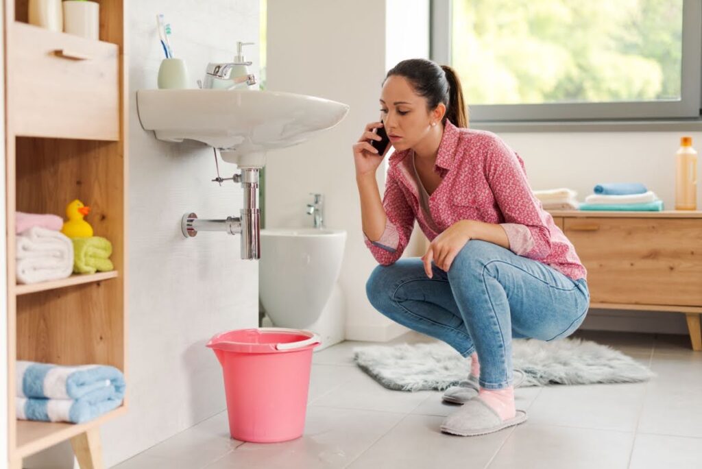 a woman kneeling next to the bathroom sink, which has a bucket under it because it has a clog that's backing up