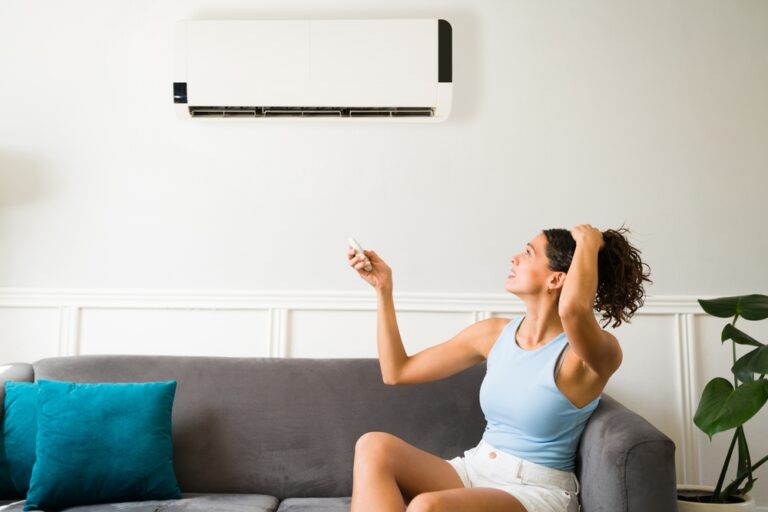 young woman sitting on the couch, holding her hair up with one hand and trying to turn on her ductless AC mini split with a remote in the other hand
