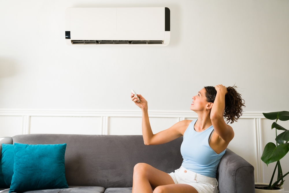 young woman sitting on the couch, holding her hair up with one hand and trying to turn on her ductless AC mini split with a remote in the other hand