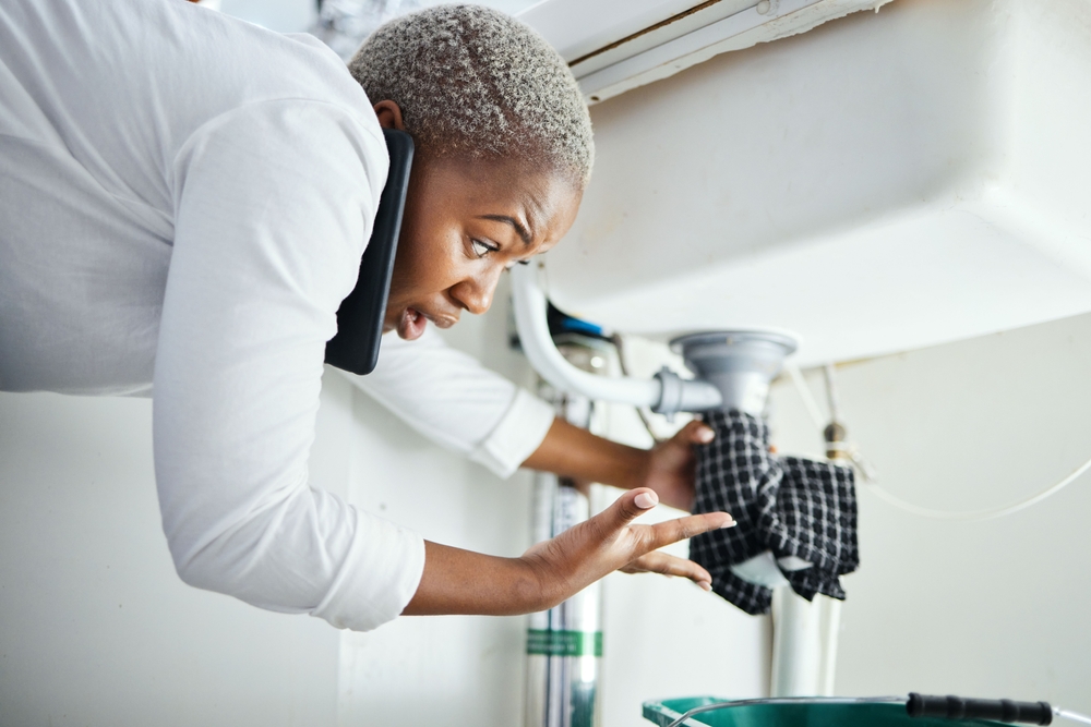 Woman, stressed, on the phone with a plumber as she holds a towel around a leaking pipe under the sink
