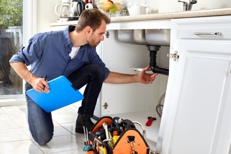 a professional plumber with clipboard in hand examining the piping under the kitchen sink during a plumbing inspection